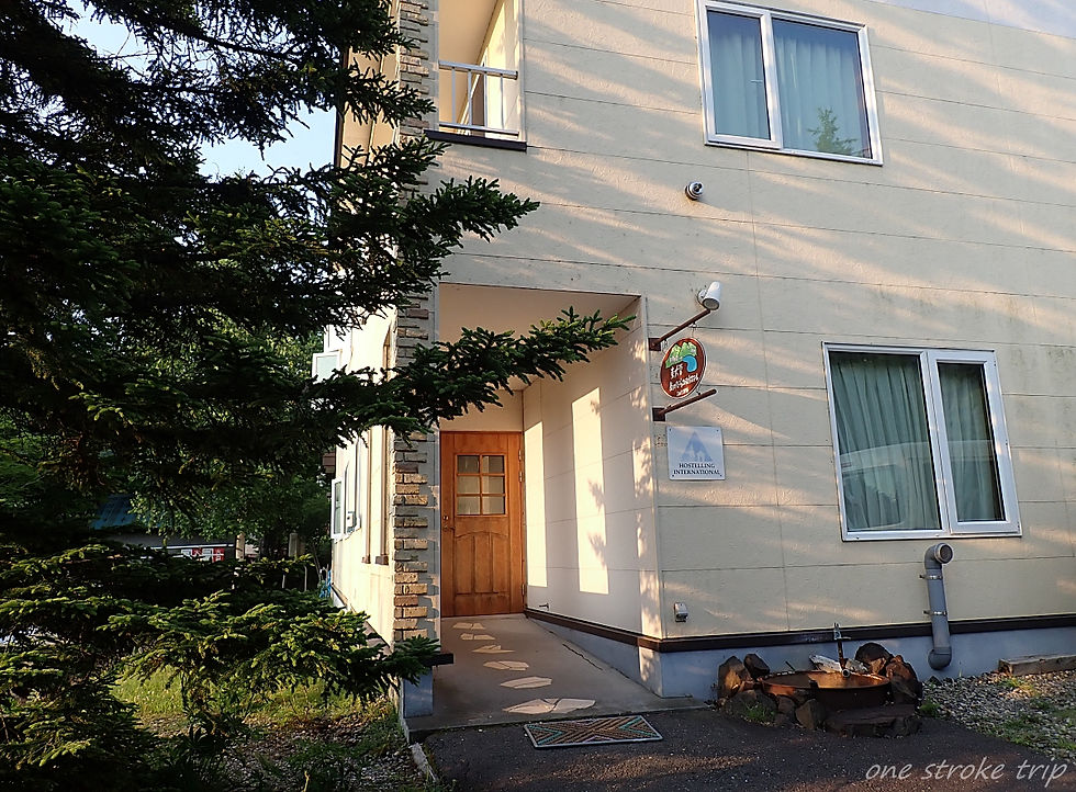 木々越しに見えるベージュの宿泊施設の外観。木製ドアと窓、壁に「HOSTELLING INTERNATIONAL」看板、夕方の柔らかな日差し。