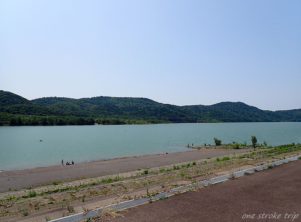 晴天の下、緑の山々に囲まれた湖畔。砂浜や階段状の岸辺に数人がたたずむ