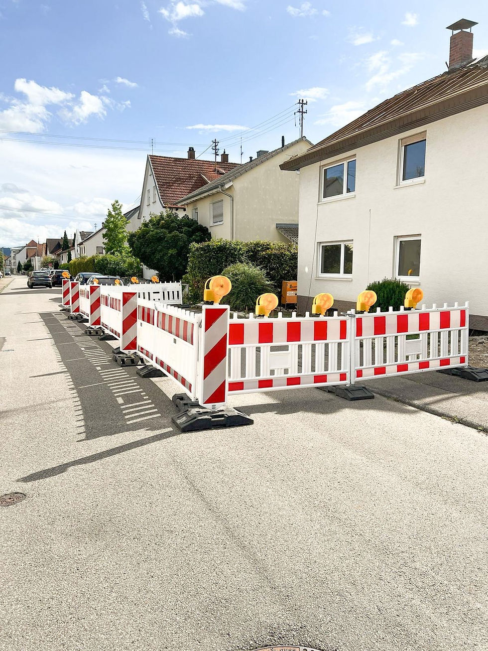 Straßensperre mit Verkehrssicherung, rote und weiße Barrieren, Häuser im Hintergrund.