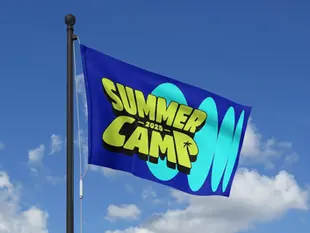 Bandeira com fundo azul vibrante exibindo o logo 'Summer Camp 2025' em amarelo com sombra preta, hasteada sob um céu claro com algumas nuvens.