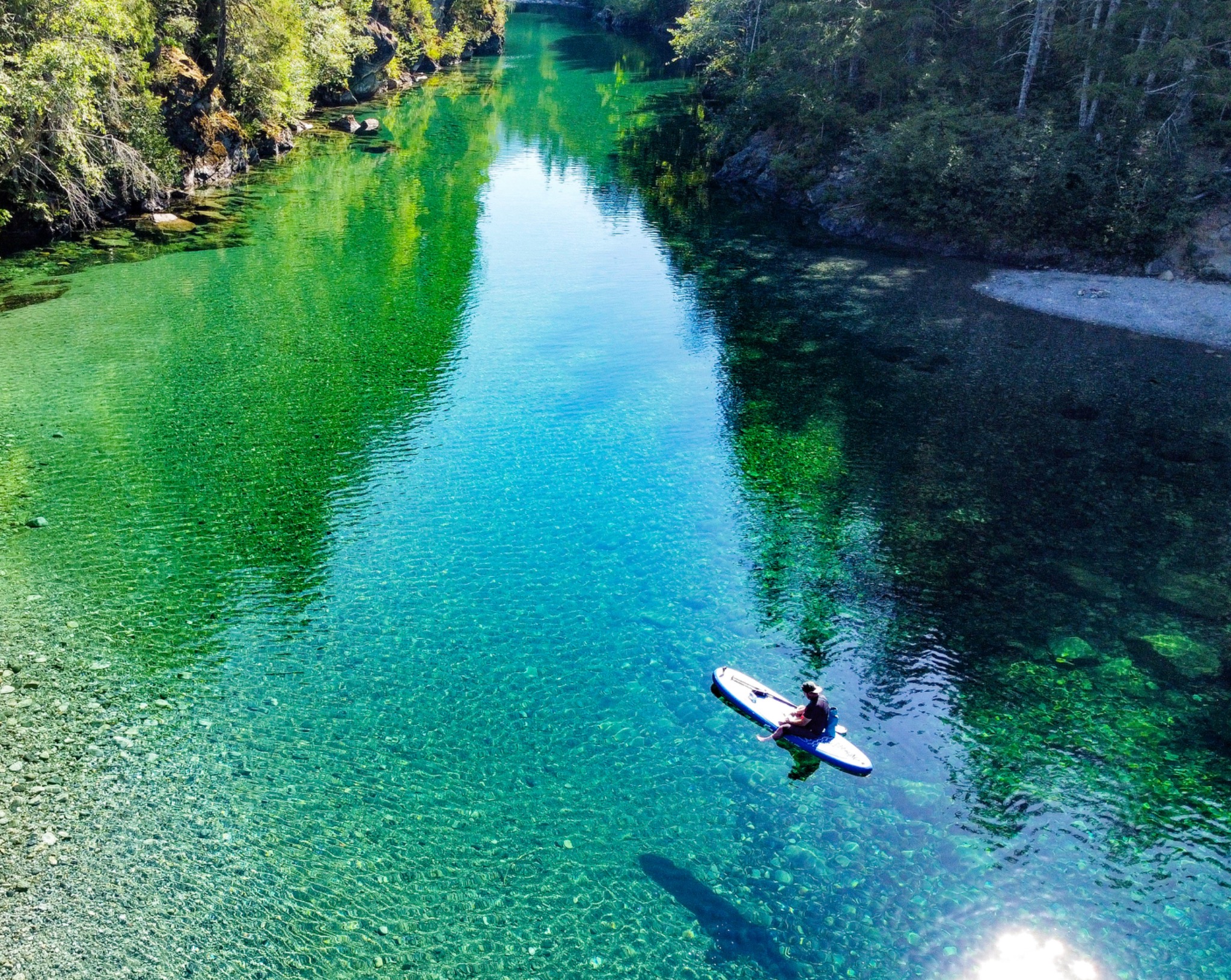 Drone photograph of a person paddleboarding through clear turquoise-green river water surrounded by forest in British Columbia