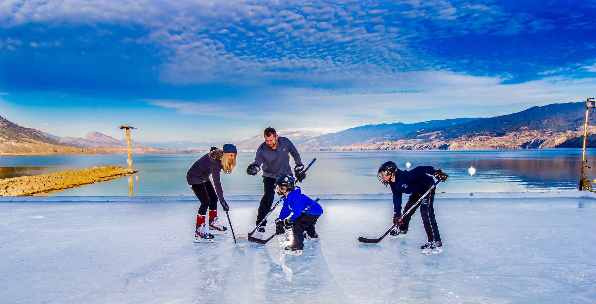 Family playing recreational ice hockey together on a frozen outdoor rink beside an Okanagan lake and surrounding mountains