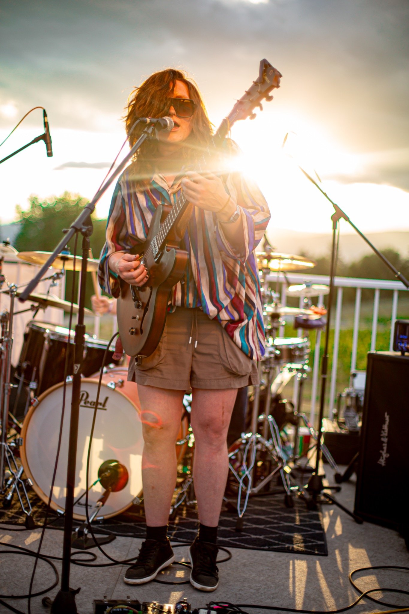 Musician in sunglasses playing guitar and singing into a microphone on an outdoor stage in warm evening sunlight