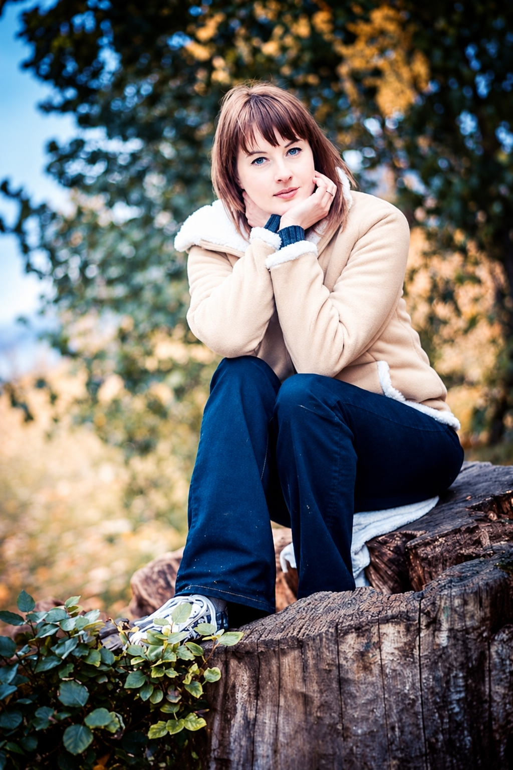 Natural light outdoor portrait of a woman in a cream jacket seated on a weathered tree stump among trees