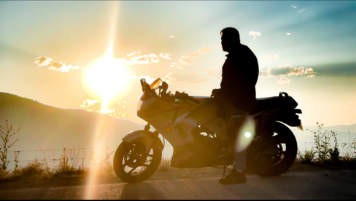 Silhouetted motorcyclist seated on a touring motorcycle beside a lake as the sun sets over distant hills