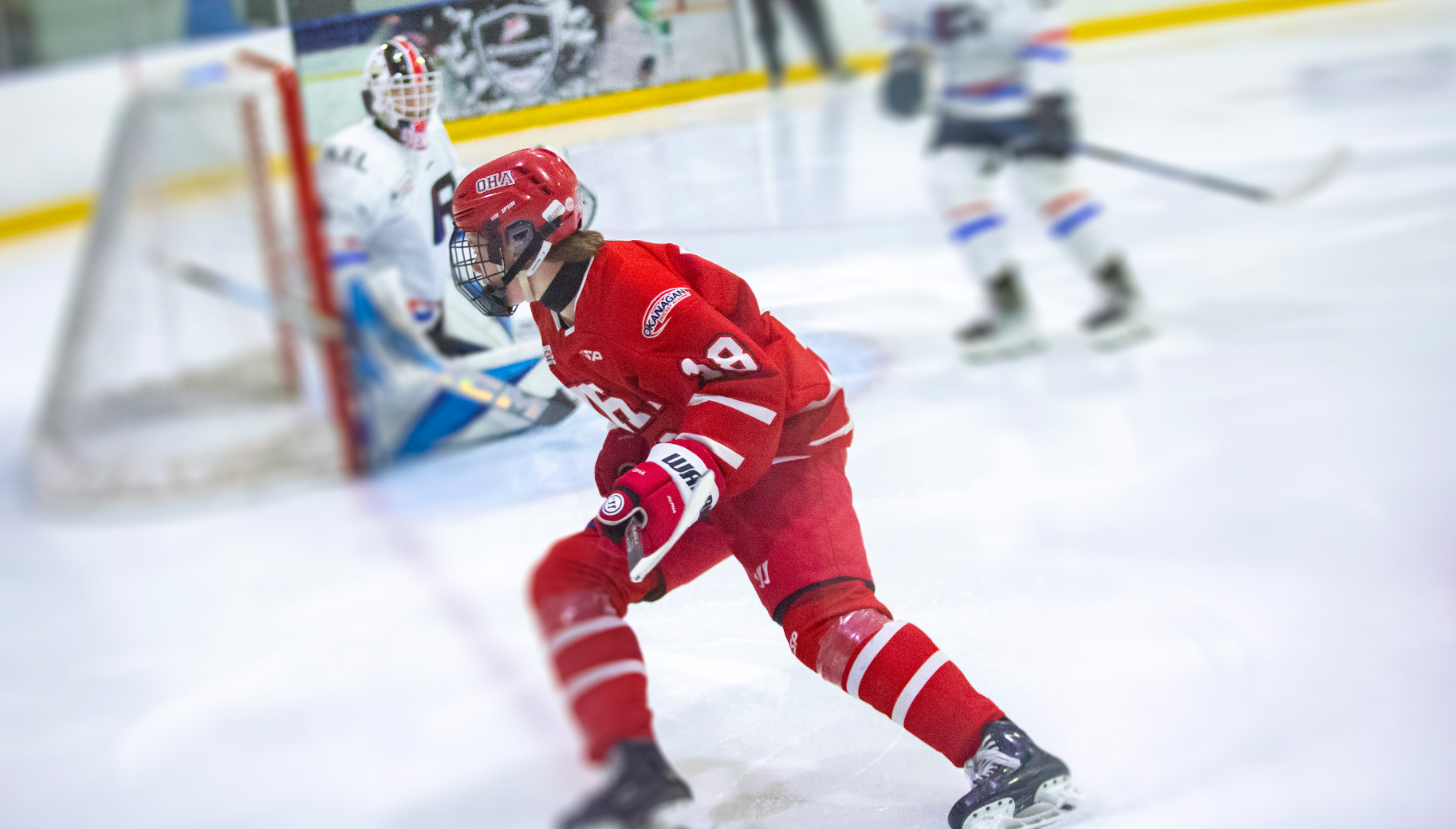 Okanagan Hockey Academy player wearing red number 8 skating with the puck near the opposition goal during a game