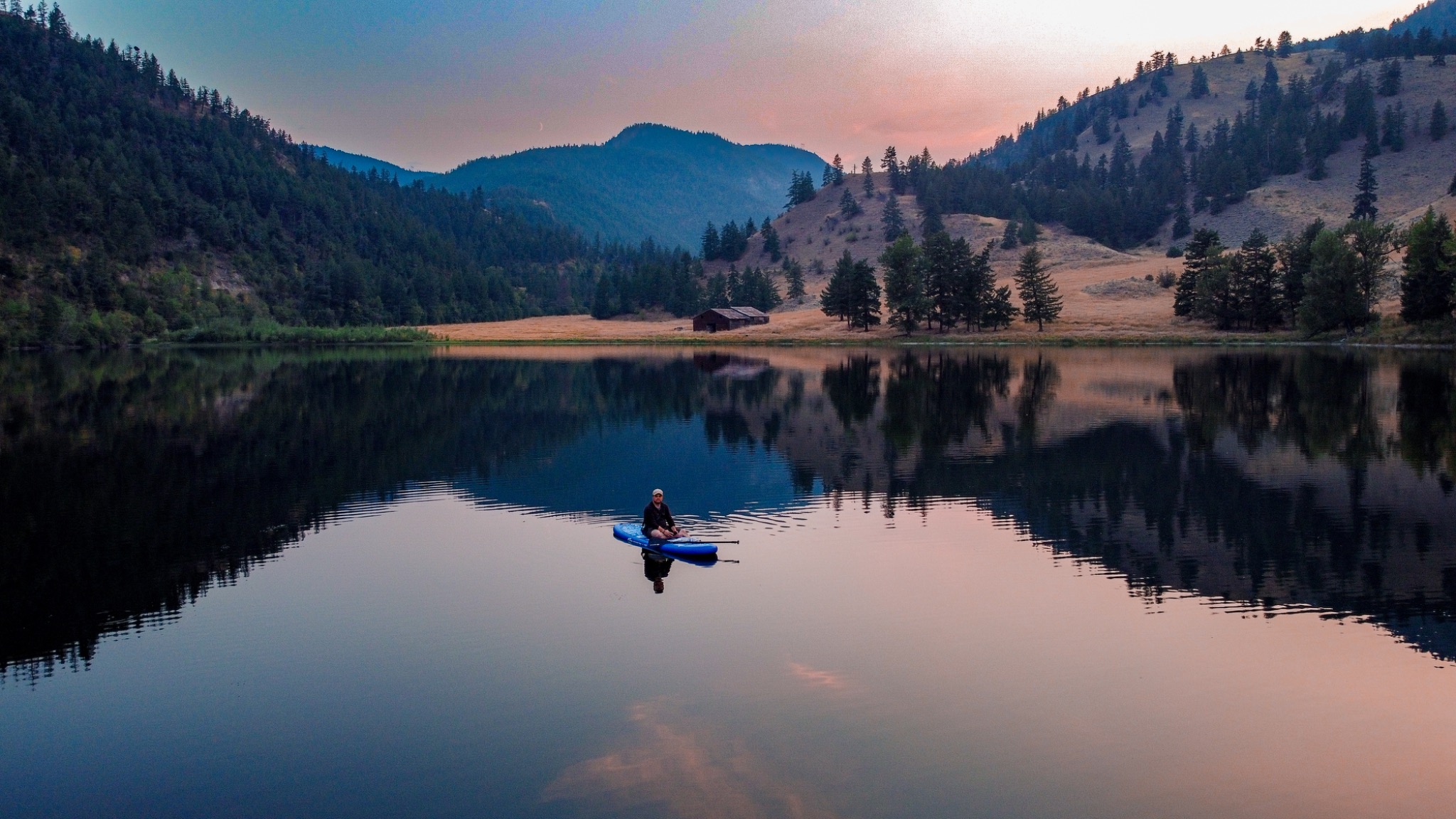 Single person sitting on a paddleboard on a mirror-still mountain lake at dusk with pink sky reflections and forested hills