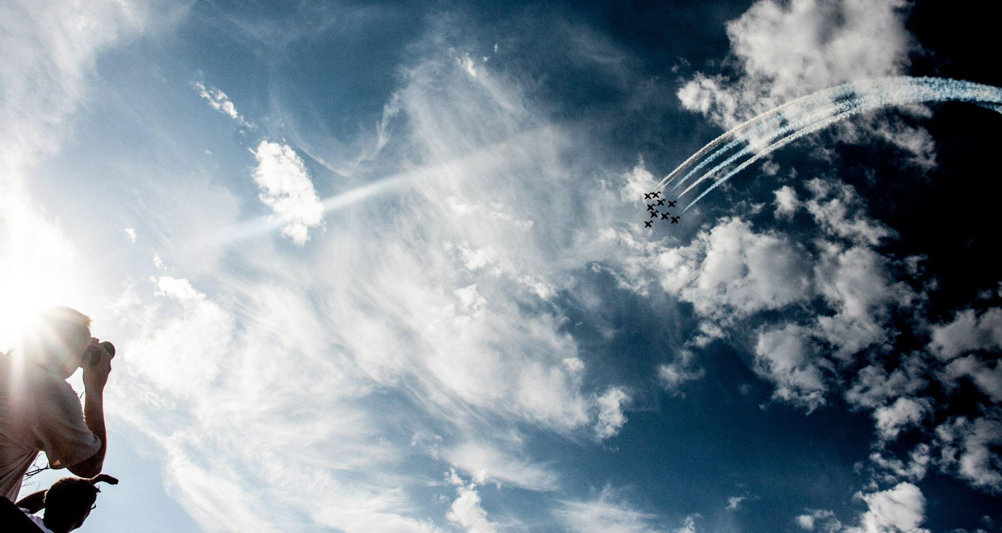 Aircraft flying in tight formation with white smoke trails curling through a dramatic blue sky as an observer photographs the display