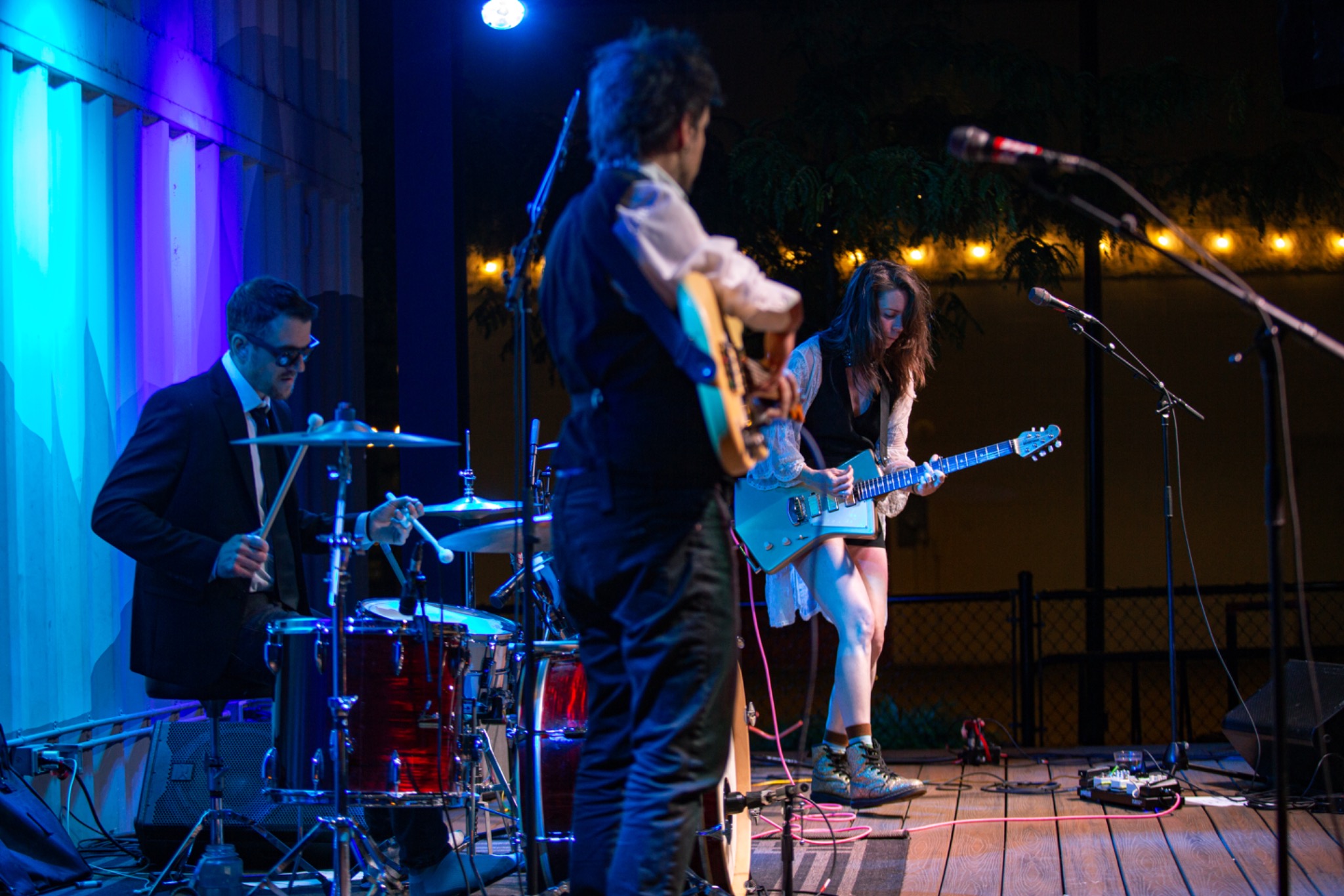 Drummer, bassist and electric guitarist performing live on a stage lit with blue and violet lights