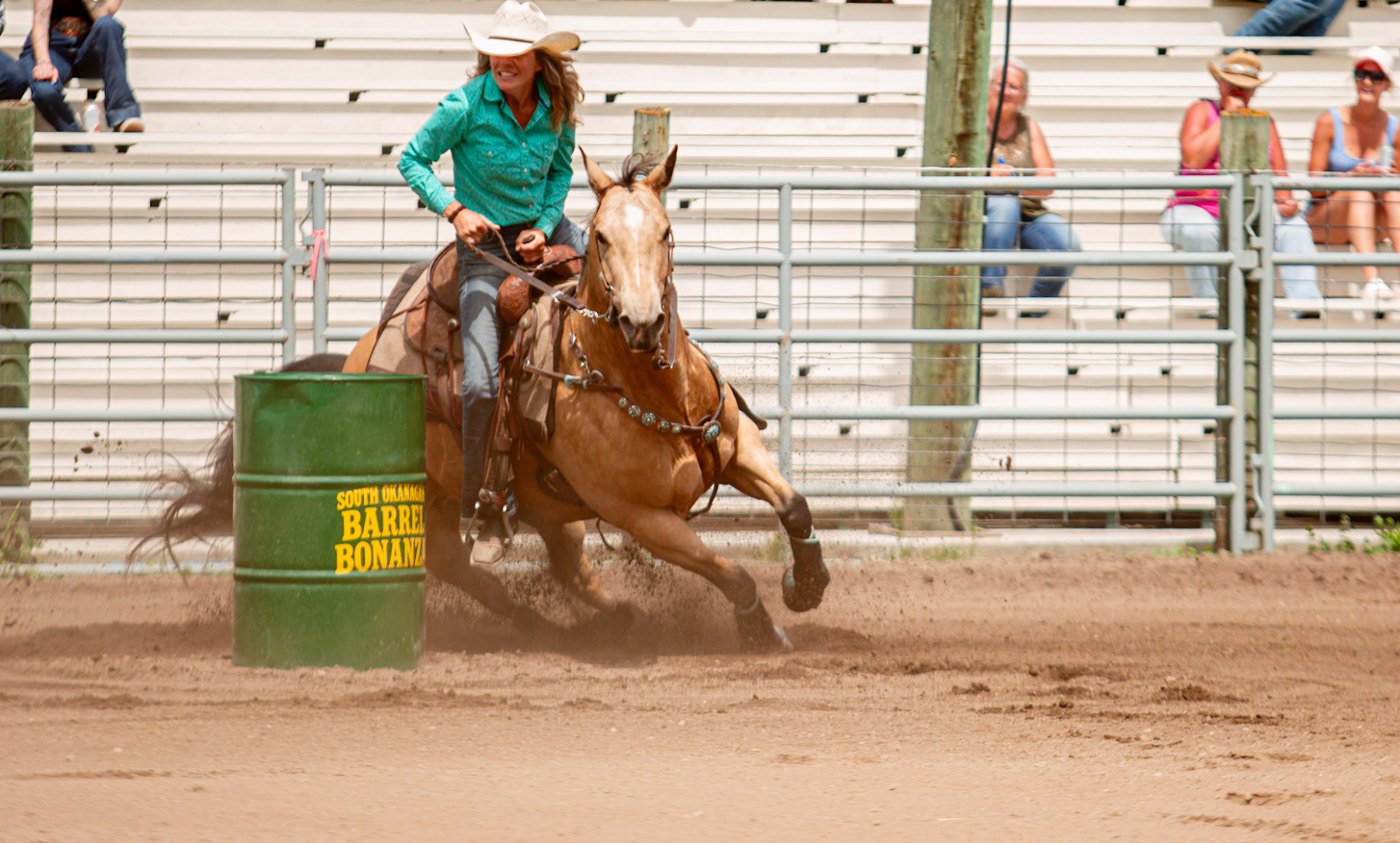 Barrel racer in a turquoise shirt guiding a brown horse around a green barrel at the South Okanagan Barrel Bonanza