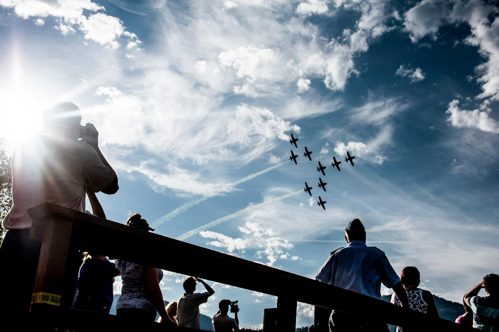 Spectators watching a synchronized aircraft formation fly overhead against a dramatic cloud-filled sky at an Okanagan air display