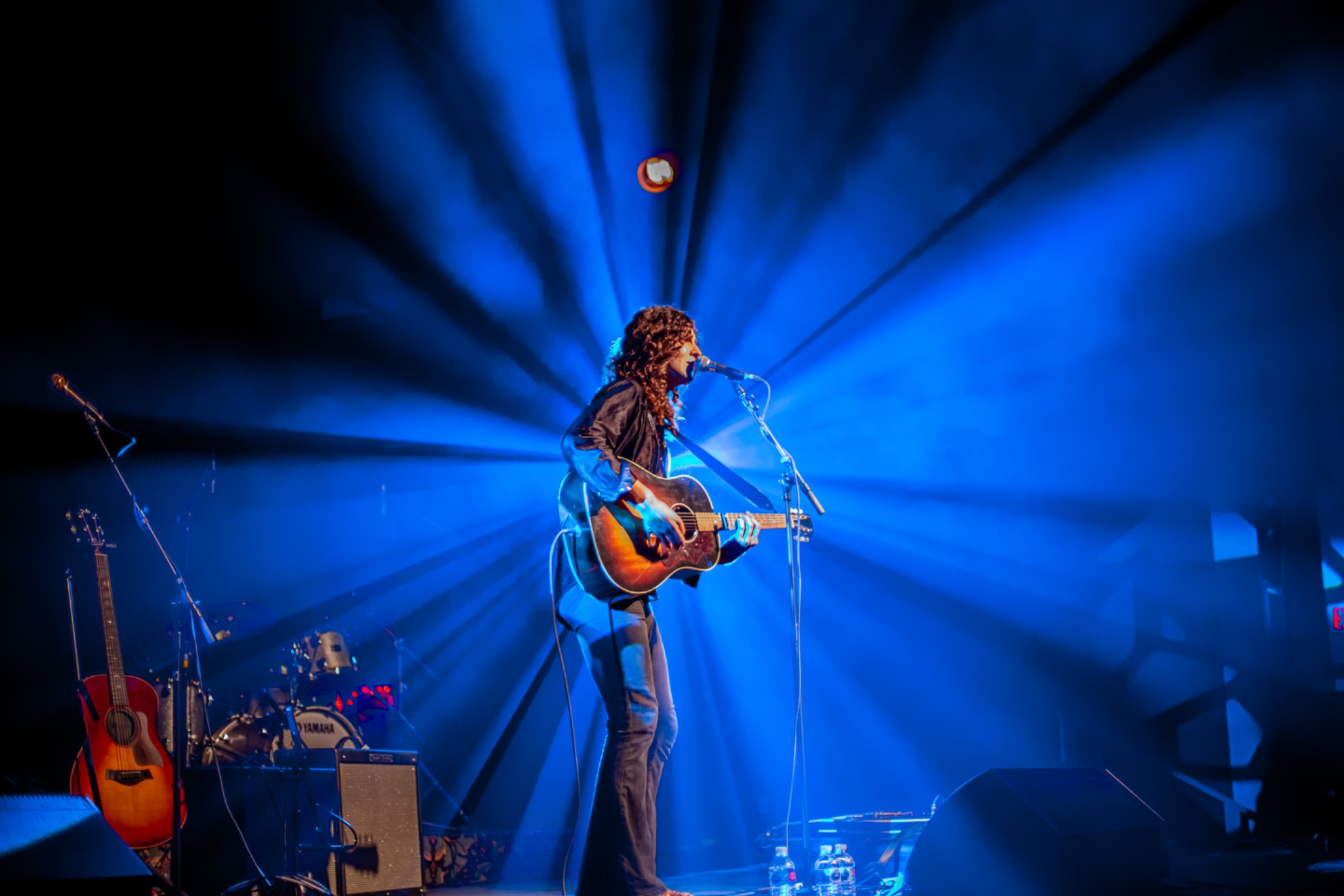 Singer-songwriter playing acoustic guitar at a microphone as vivid blue spotlights radiate across the stage