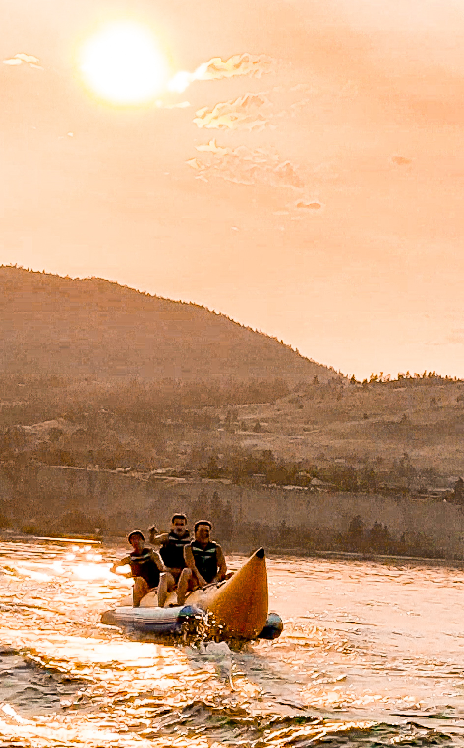 Three riders on an inflatable banana boat on sunlit lake water at golden hour with mountains behind them