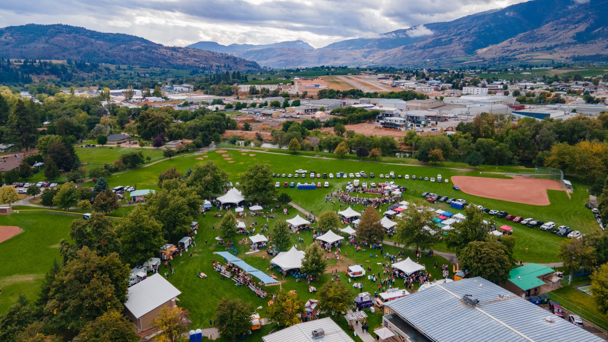 High aerial drone photograph of a community festival in a green park in Oliver, BC, with event tents, gathering crowds, fields and surrounding mountains