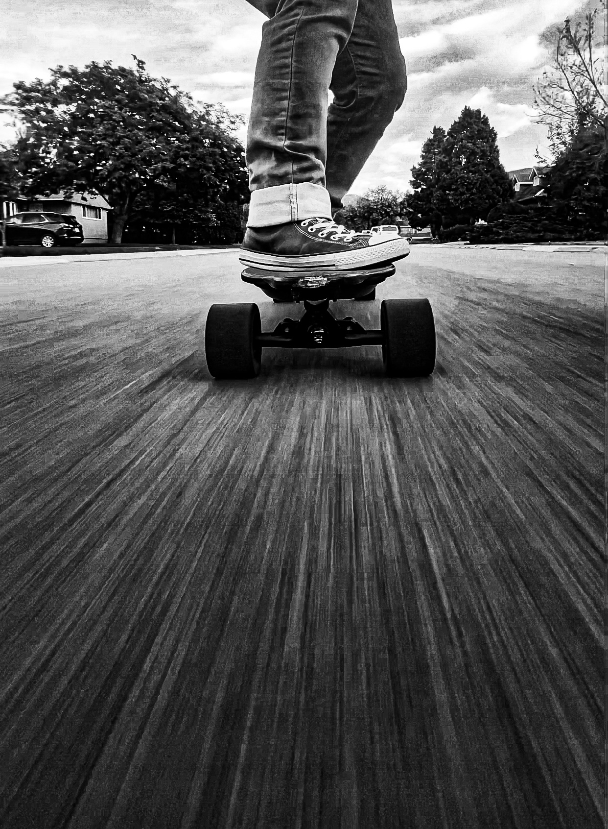 Black-and-white low-angle view of a sneaker on an electric longboard rolling along a paved residential street