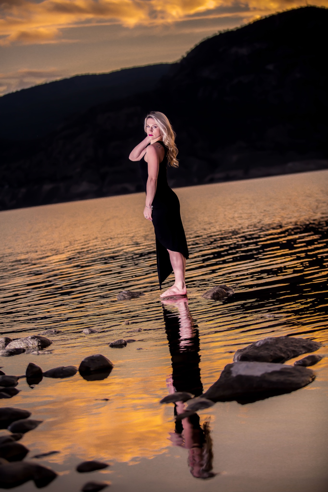 Woman in a black dress standing in shallow lake water at sunset with her reflection