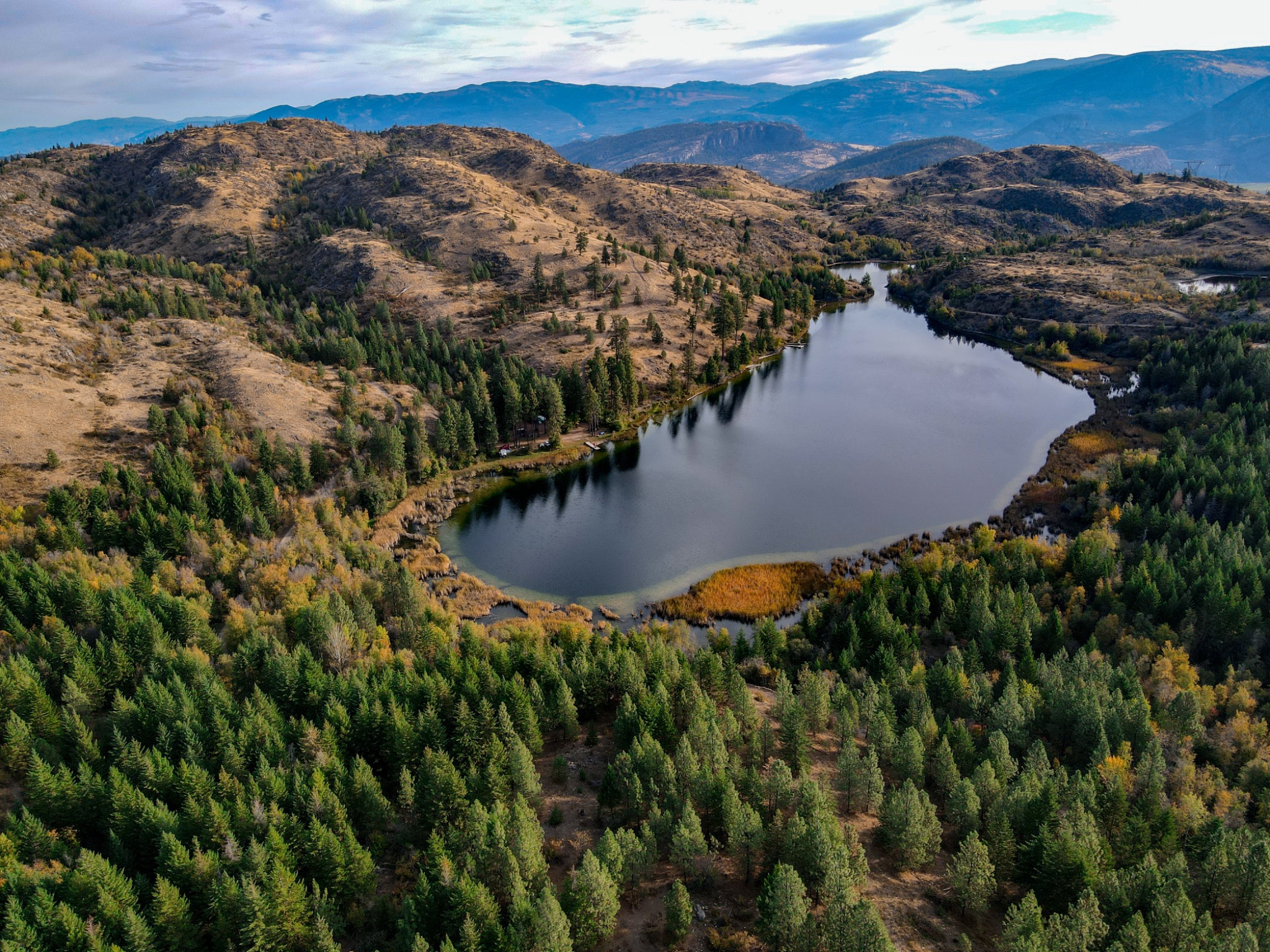 Aerial view of a forest-lined mountain lake framed by dry rolling hills and distant Okanagan-style mountain ridges