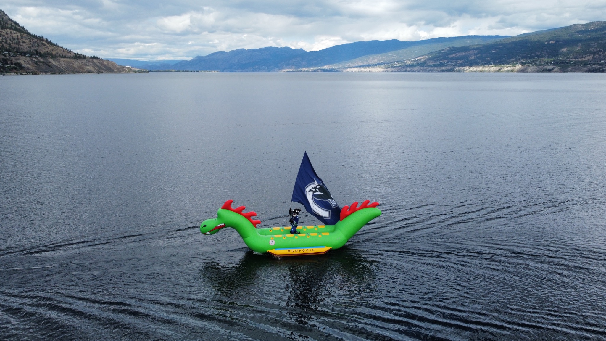 Green Ogopogo-shaped inflatable boat with flag floating on Okanagan Lake beneath distant mountains and clouds