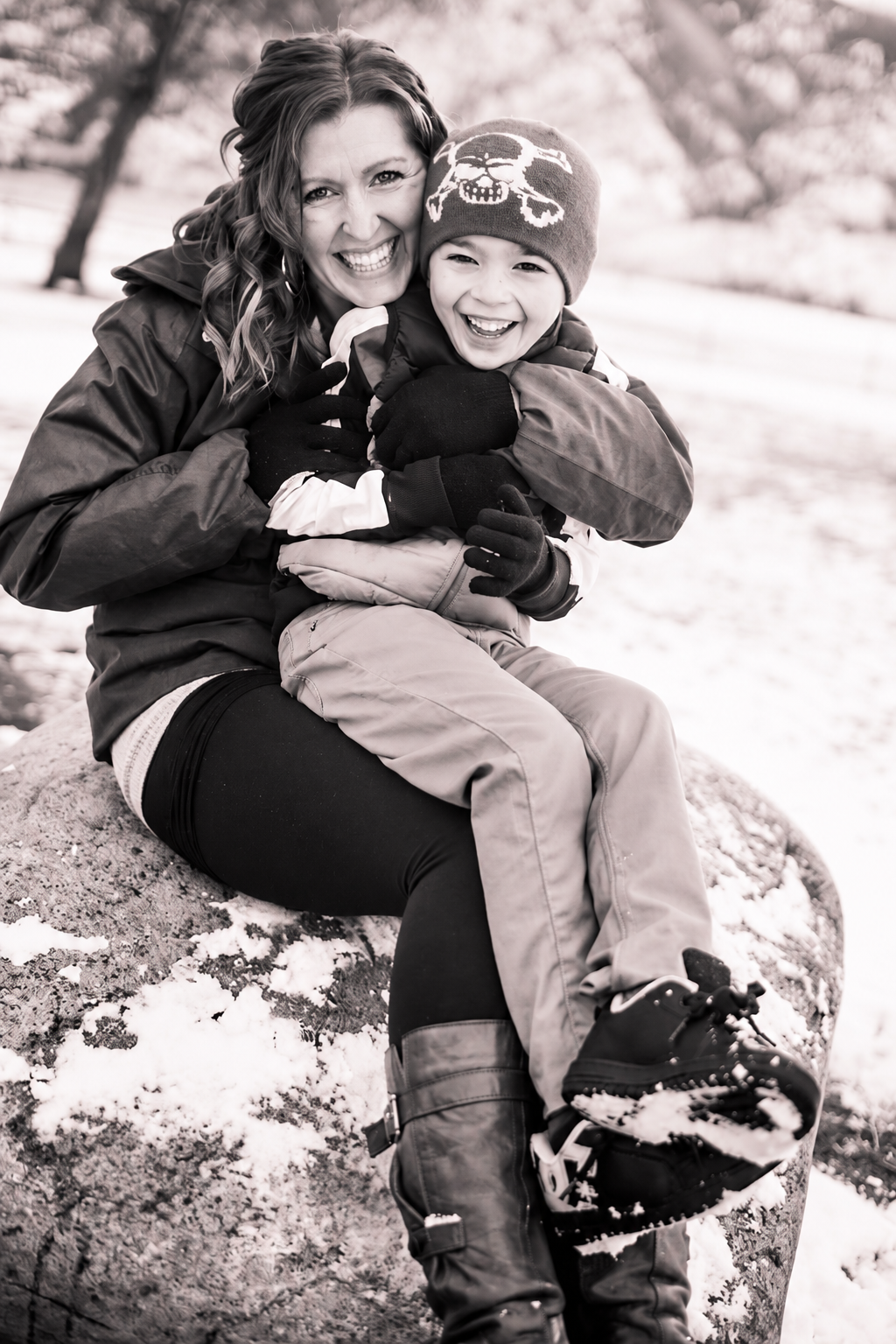 Candid black-and-white family photo of a laughing mother and son hugging on a snow-covered boulder