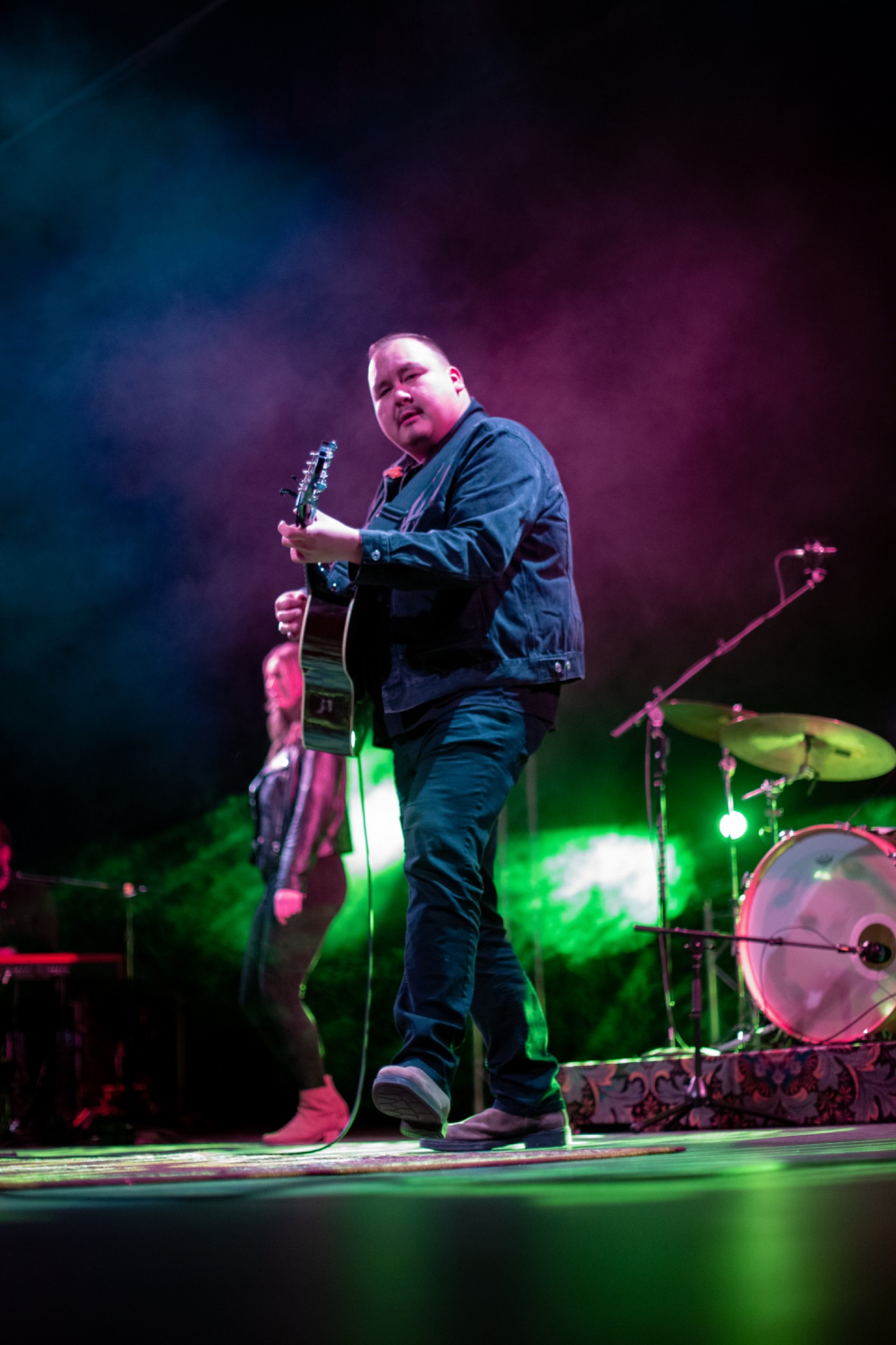 Guitarist performing on stage in front of a drum kit with magenta and green lighting and theatrical haze