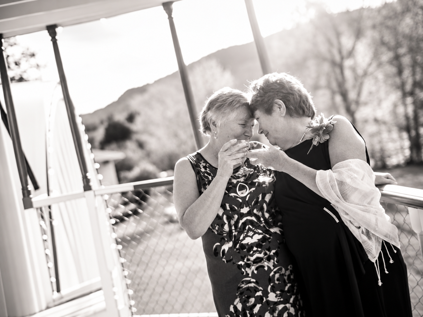 Candid black-and-white photograph of two women smiling together and raising glasses on a sunlit porch