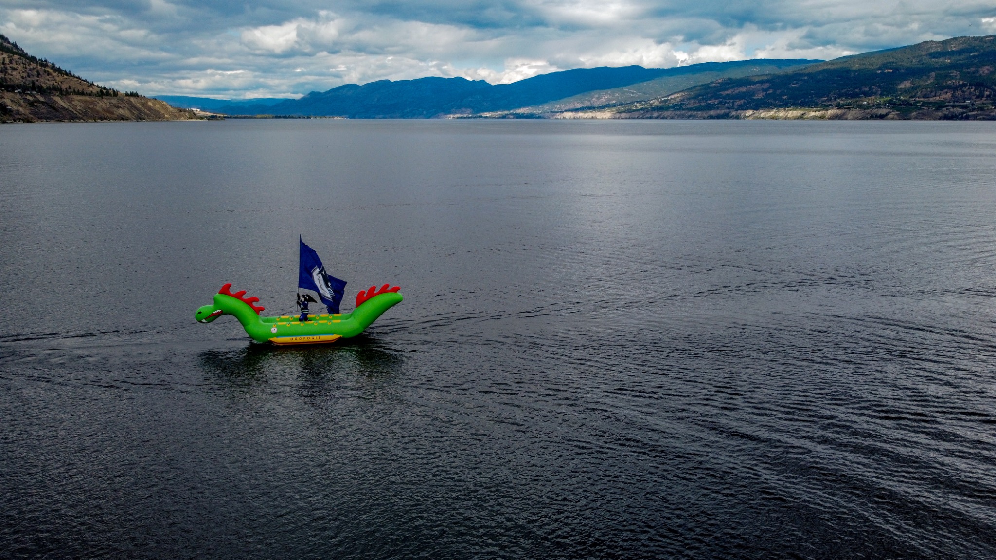 Wide aerial view of a bright green Ogopogo inflatable boat on a large lake surrounded by mountains beneath a cloudy sky