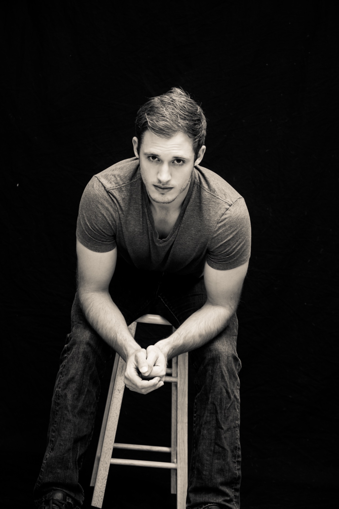 Black-and-white creative portrait of a man sitting forward on a stool against a dark background