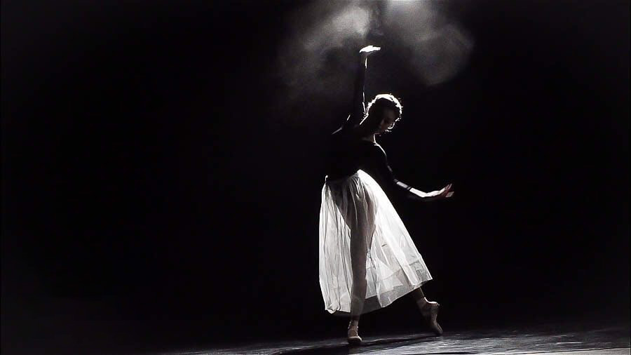 Silhouetted ballet dancer in a white flowing skirt reaching toward a spotlight on a dark theatre stage