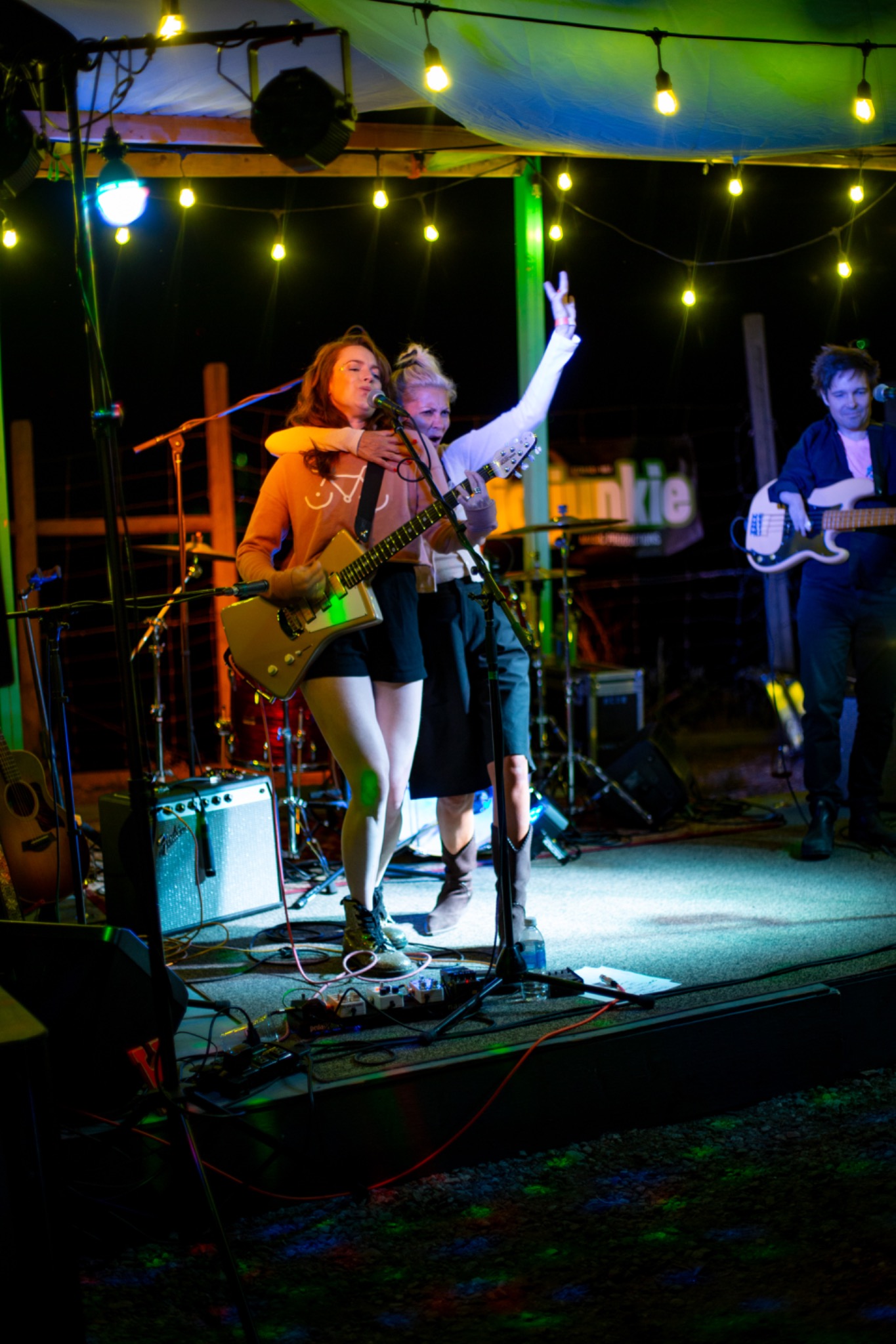 Outdoor live band performance under colourful stage lighting and string lights, with a guitarist and two musicians sharing a lively moment onstage in the Okanagan