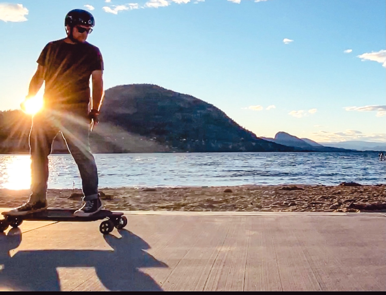 Helmeted rider standing on an electric longboard on a lakeside paved path in warm late-afternoon light