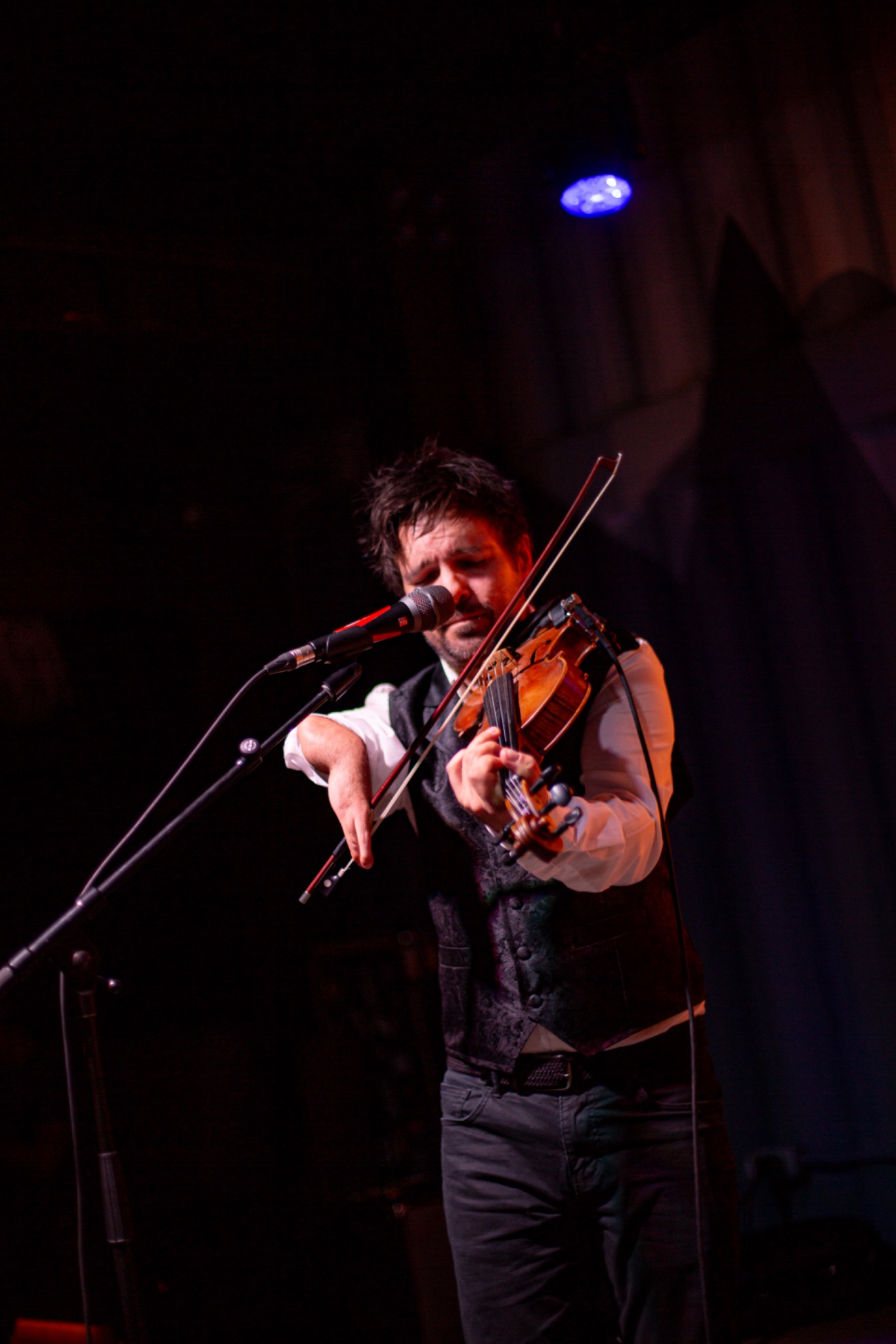 Violinist playing and singing into a microphone on a dark stage with warm red performance lighting