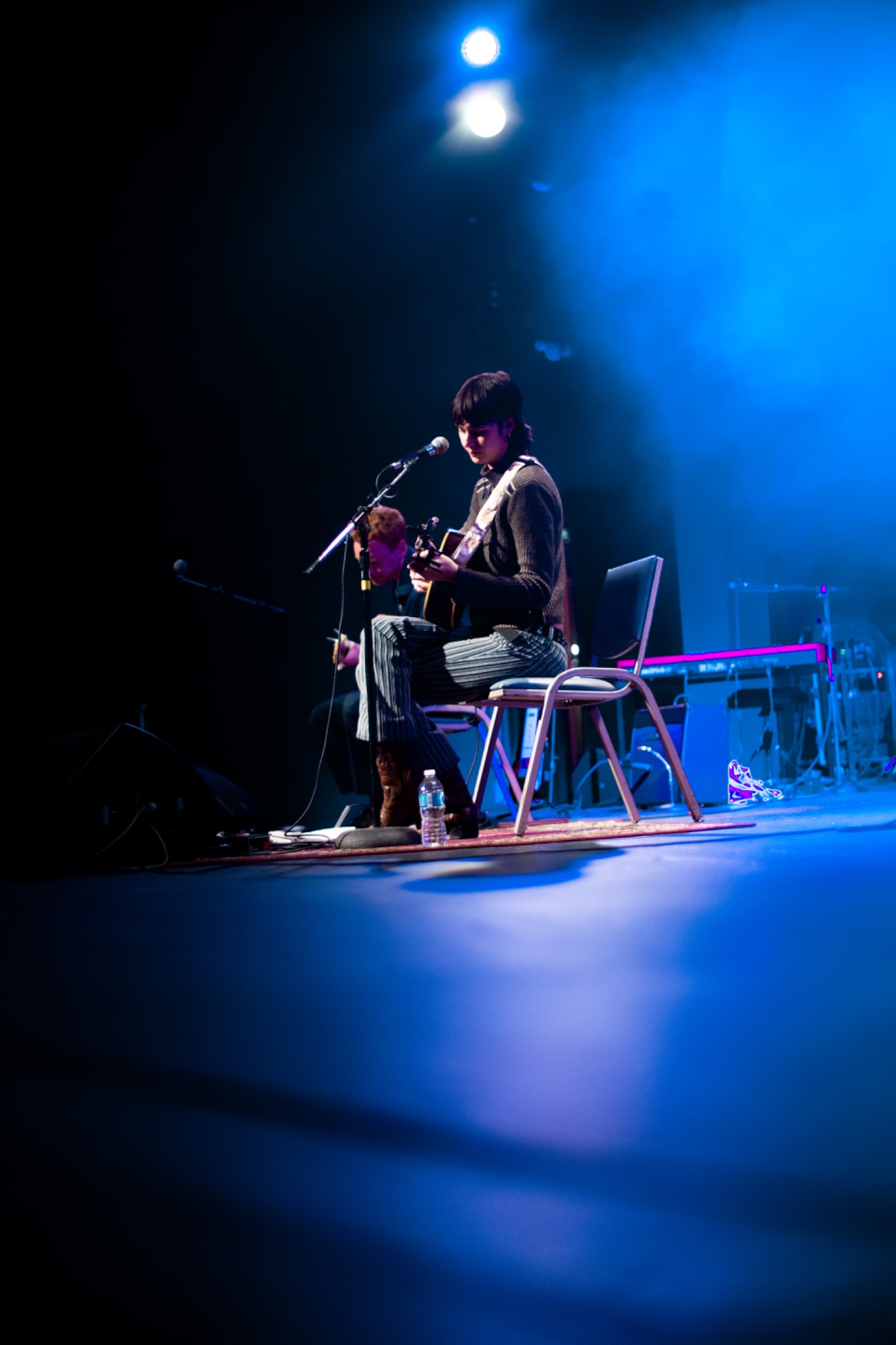 Seated performer playing guitar and singing into a microphone on a softly blue-lit stage