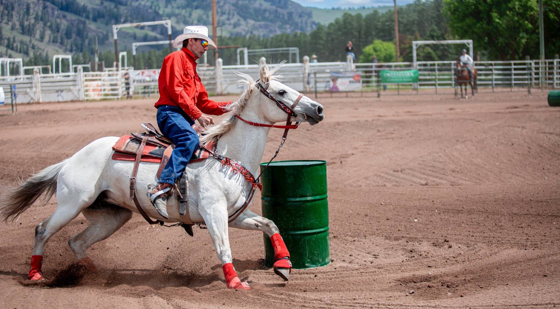 Rider wearing a red shirt and cowboy hat racing a white horse past a green barrel during a rodeo event