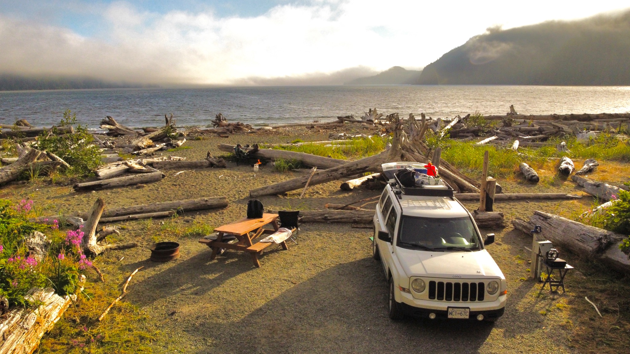 White Jeep parked beside a driftwood-covered coastal campsite in British Columbia with the water and distant headlands behind it