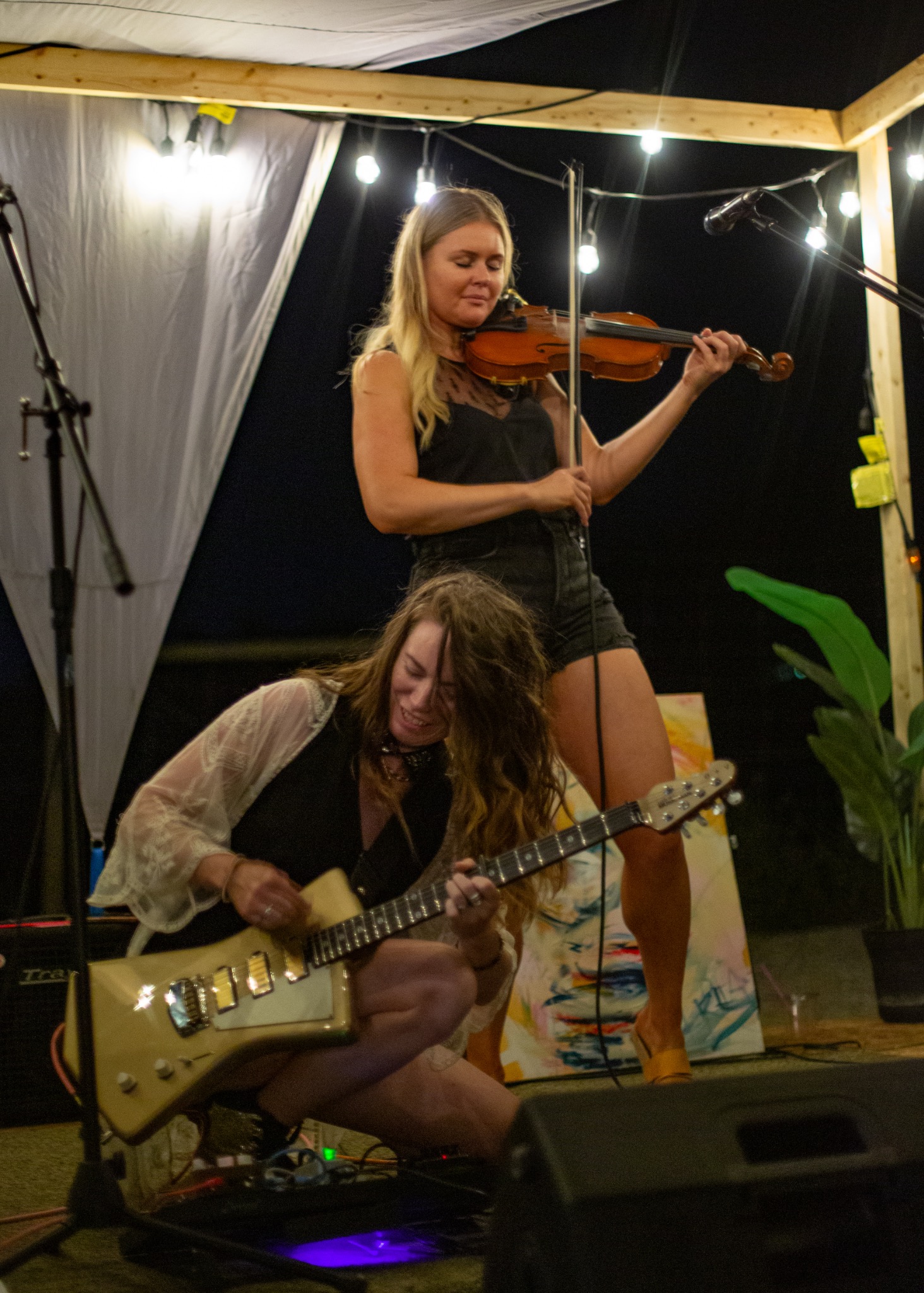 Live music duo performing onstage at night, with one musician playing electric guitar and another playing violin beneath string lights