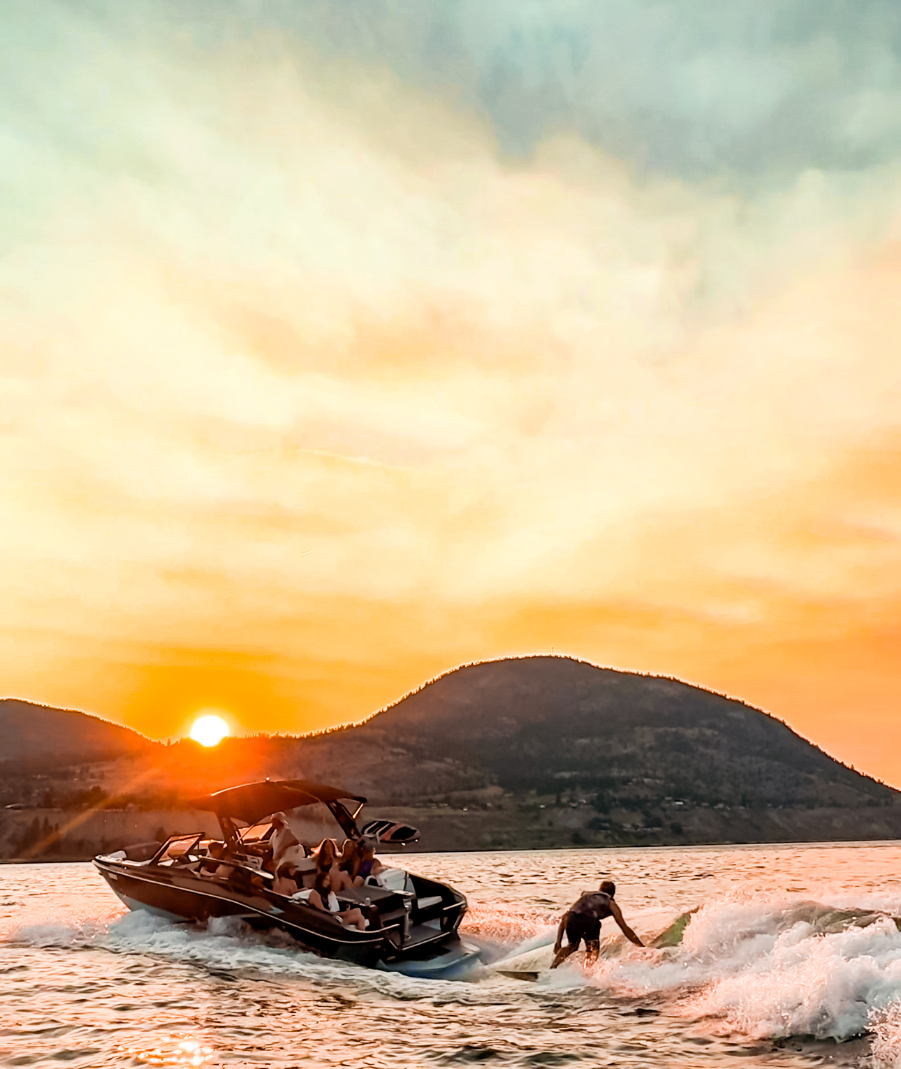 Boat towing a wakesurfer across a lake at sunset, with golden light on the water and a silhouetted ridge behind