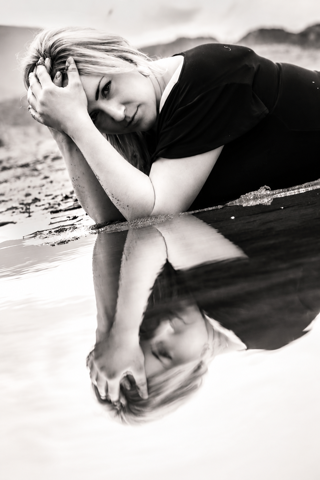 Creative black-and-white waterside portrait of a woman resting by still water with her face reflected below