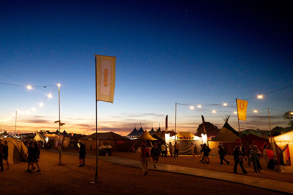 Festival early morning scene at sunrise with tents, flags, and string lights, as people walk across the site under a brightening blue and orange sky