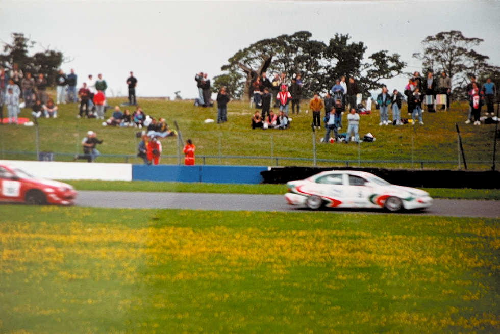 Touring cars race past spectators on a grassy bank at a motorsport circuit, Donington Park, with motion blur capturing the speed.