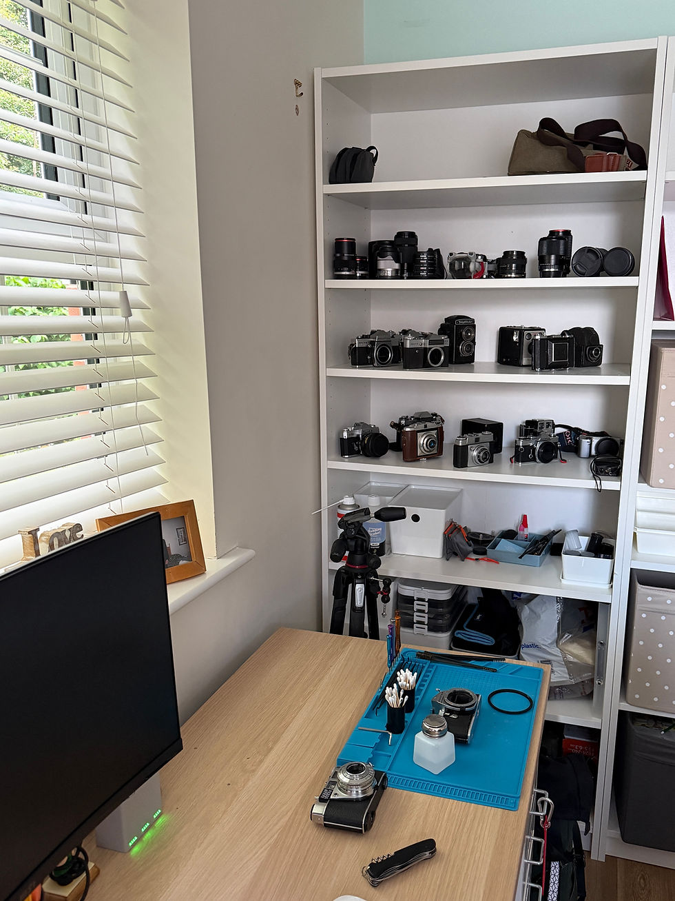Home photography workspace featuring a shelving unit filled with vintage cameras and lenses, a tripod, and a tidy desk setup with cleaning tools and film cameras ready for maintenance beside a computer monitor.