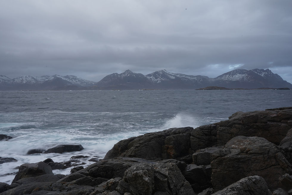 Moody long exposure of waves crashing on rocks along the Norway coast, with snow-dusted mountains in the distance and dramatic storm clouds overhead.