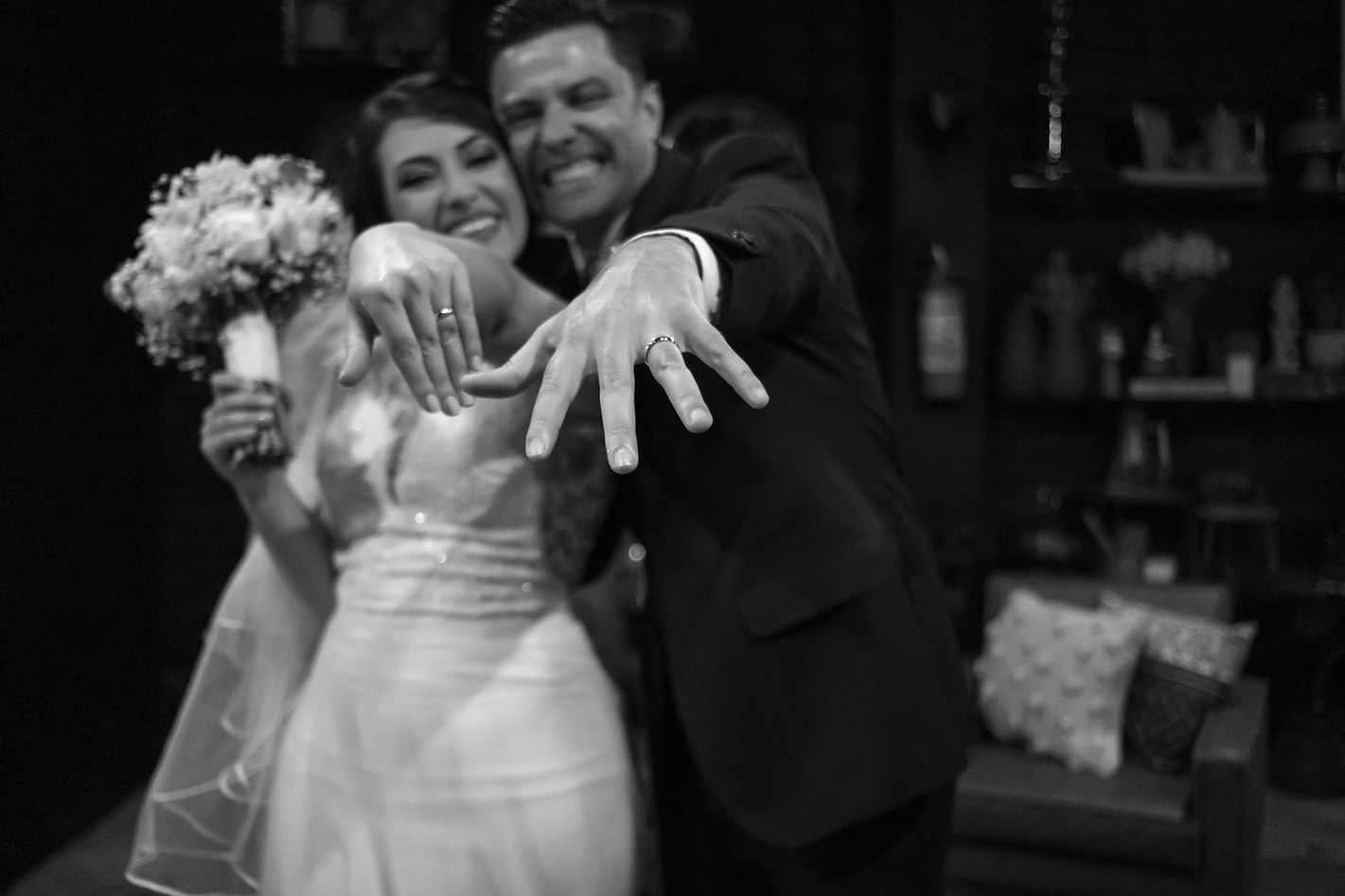 Casal sorri feliz em uma foto em preto e branco, mostrando suas alianças, em celebração do dia do seu casamento