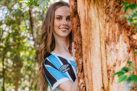 Moça jovem sorrindo próximo à arvore, com camisa do seu time favorito