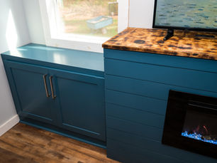 Blue cabinet with silver handles next to a fireplace and wooden countertop. Window view of green outdoor setting. Cozy and modern ambiance.