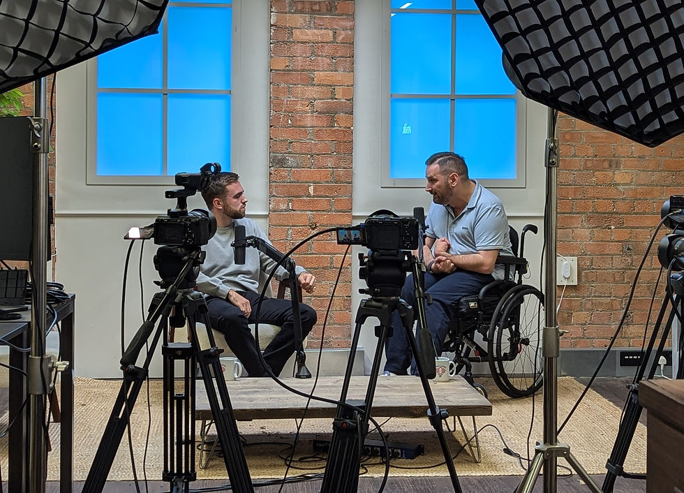 Image description. Josh and Adam chatting in the background of the podcast studio with camera and lighting equipment in the foreground.