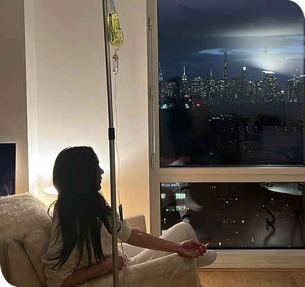 Woman sitting on a couch receiving an IV drip in a cozy apartment, with a large window view of the New York City skyline at n
