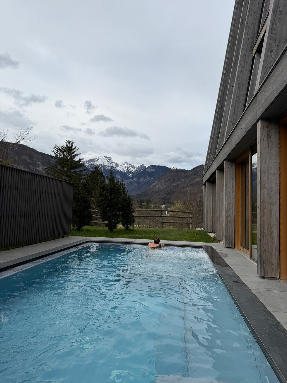 Person im Pool vor modernem Holzgebäude. Berge im Hintergrund, bewölkter Himmel. Entspannte Stimmung.