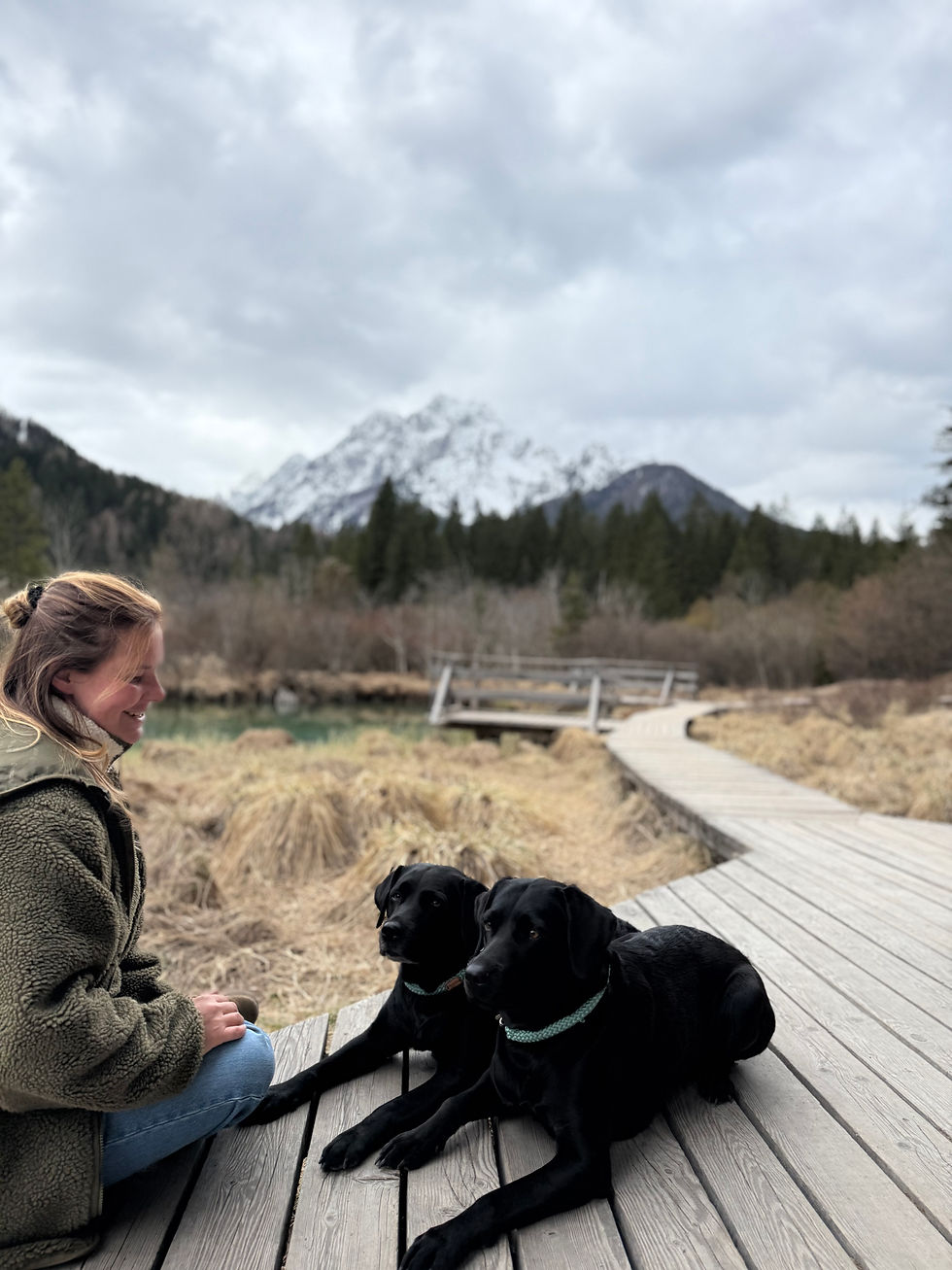 Frau sitzt lächelnd auf einem Holzsteg neben zwei schwarzen Hunden. Im Hintergrund schneebedeckte Berge und bewölkter Himmel.