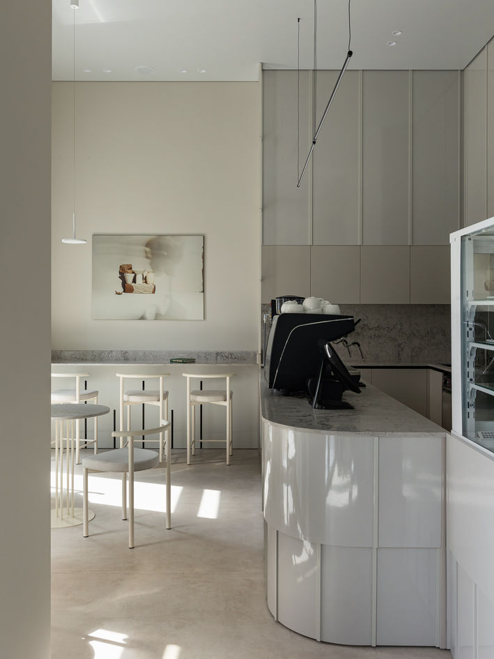 Modern minimalist café interior with white round tables, textured cream chairs, hanging pendant lights, and a sleek black coffee bar lined with cups.