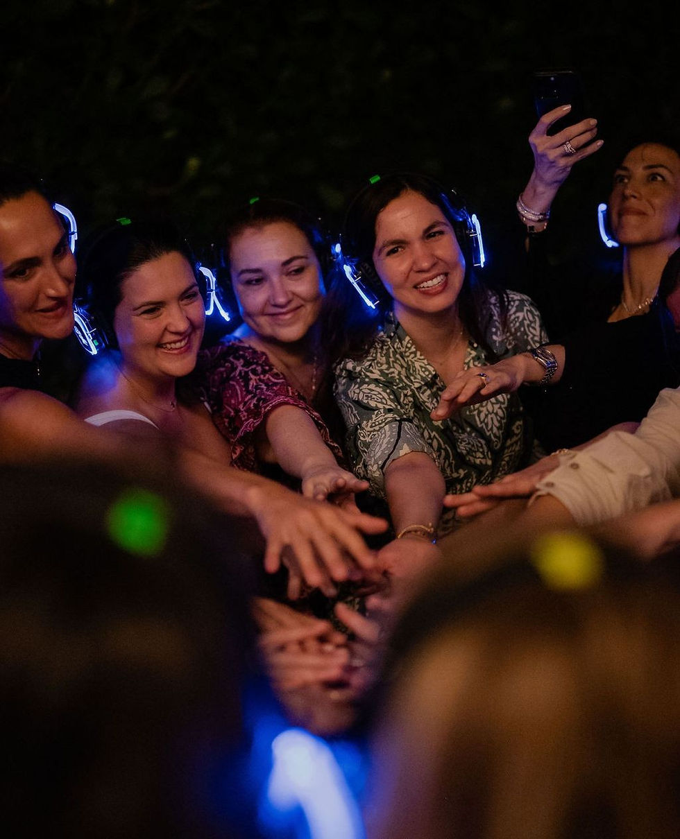 A group of people wearing glowing wireless headphones smile and reach their hands together during a Be You Disco night event, sharing a moment of joy and unity on the dance floor.